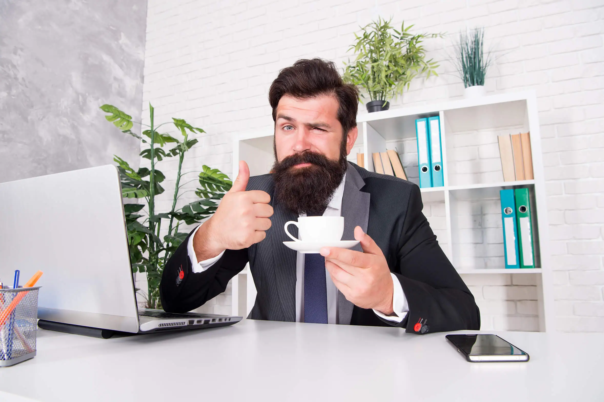 Hipster en costume, assis à un bureau, pouce levé et tenant une tasse de café, en faisant un clin d'oeil