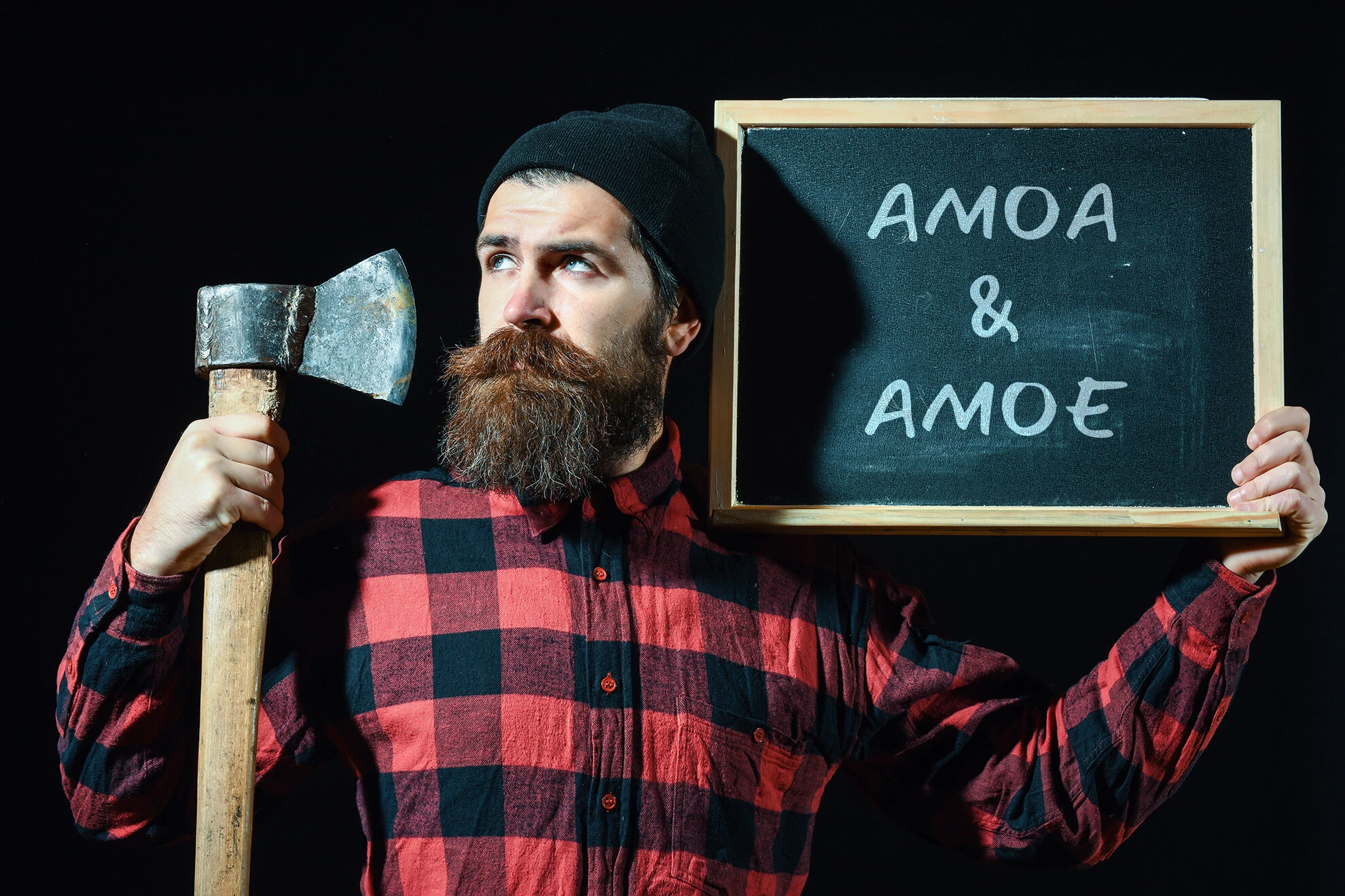 Homme barbu avec une chemise rouge et noire, un bonnet noire , tenant une hache et une grande ardoise indiquant AMOA & AMOE